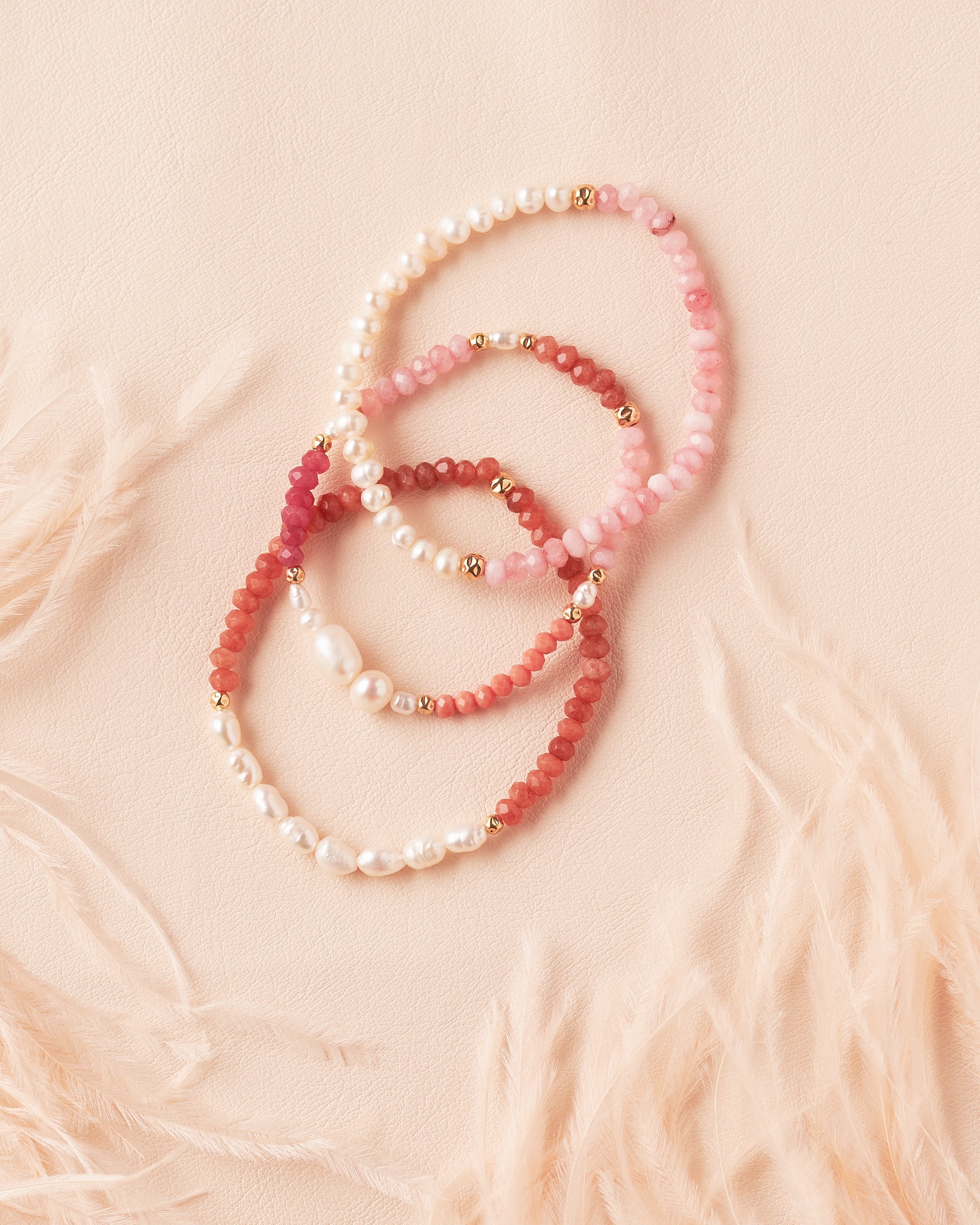 Three semiprecious and pearl bracelets in pink, brown and ombre and gold on a soft ivory fabric background